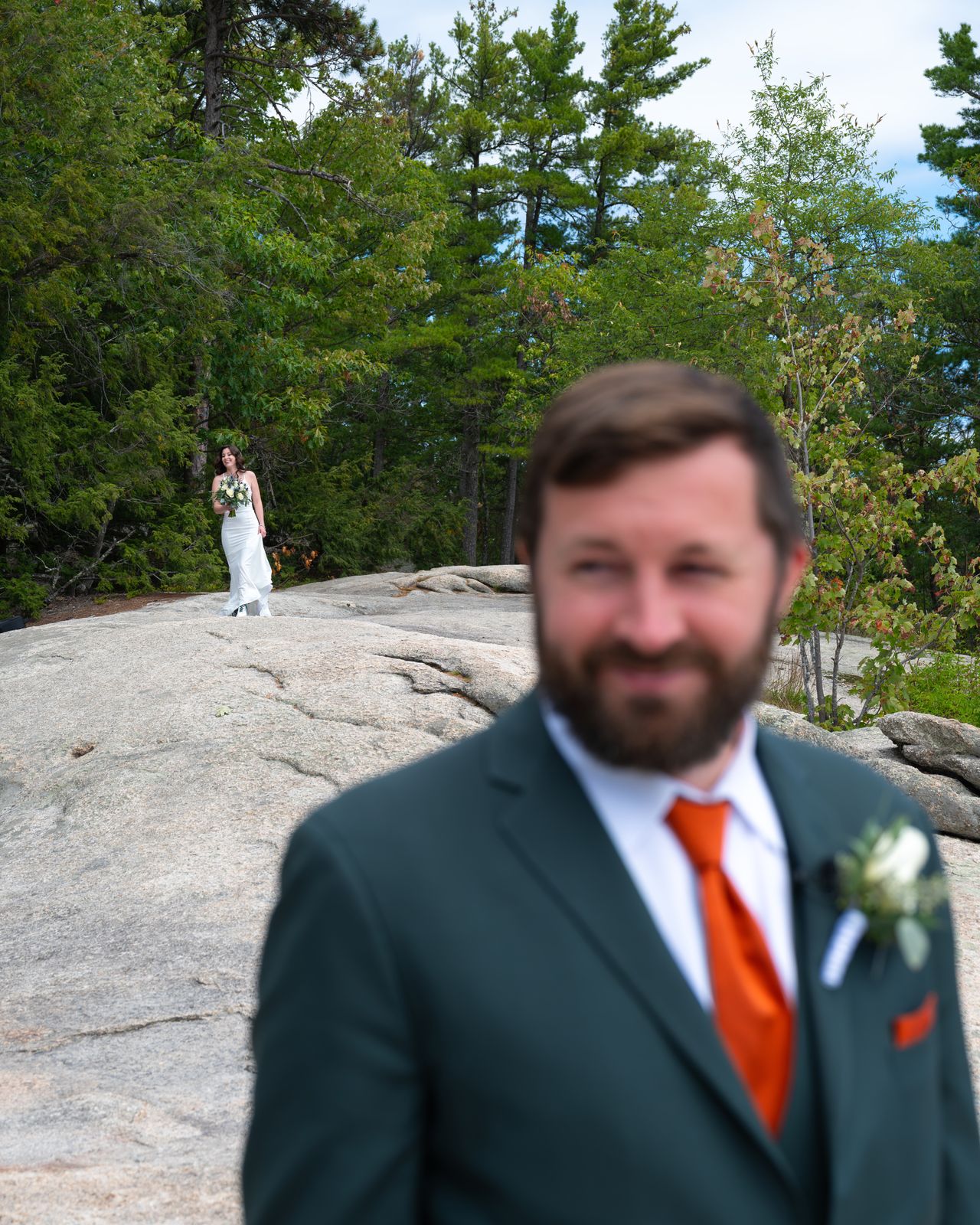 Groom in a green suit with an orange tie on Cathedral Ledge as the bride stands on the granite behind him