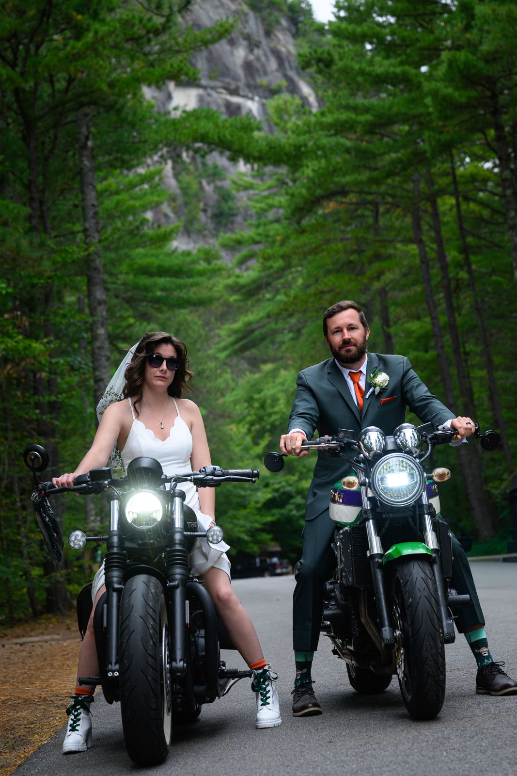 Bride in a wedding dress and veil and groom in a green suit seated on their motorcycles below Cathedral Ledge